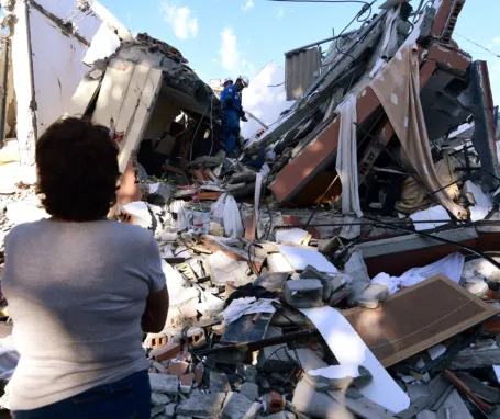 A woman looks at rescuers searching for survivors amid the rubble of a collapsed building after an earthquake in Pereira, Colombia on August 10, 2026. A magnitude 7.4 earthquake struck Colombia on August 10, 2026, and killed at least 111 people, damaged more than 1,500 buildings, and left an unknown number of people buried under rubble. The government declared a state of emergency to attend to the quake that shook several cities across the coffee-producing region, the Pacific coast, and neighboring Latin American countries, sending terrified residents racing into the street. (Photo by Jaime SALDARRIAGA / AFP)
