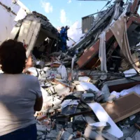A woman looks at rescuers searching for survivors amid the rubble of a collapsed building after an earthquake in Pereira, Colombia on August 10, 2026. A magnitude 7.4 earthquake struck Colombia on August 10, 2026, and killed at least 111 people, damaged more than 1,500 buildings, and left an unknown number of people buried under rubble. The government declared a state of emergency to attend to the quake that shook several cities across the coffee-producing region, the Pacific coast, and neighboring Latin American countries, sending terrified residents racing into the street. (Photo by Jaime SALDARRIAGA / AFP)